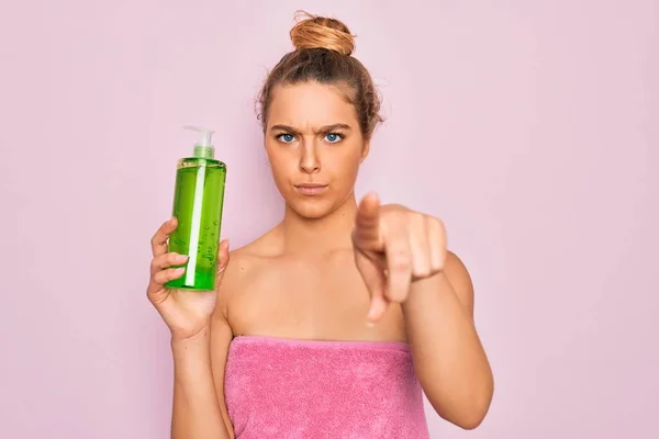 Beautiful blonde woman with blue eyes wearing towel shower after bath holding aloe vera gel pointing with finger to the camera and to you, hand sign, positive and confident gesture from the front