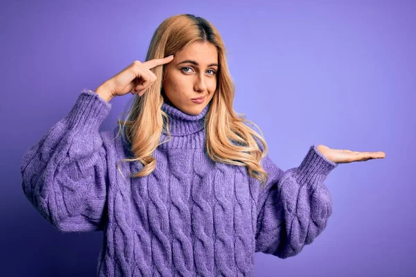 紫色の背景にカジュアルなタートルネックセーターを着た若い美しいブロンドの女性が混乱し コピースペースを示し 額に指を指すオープン手のひらに悩まされています 考えてみて — ストック写真