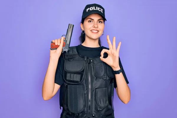 Young police woman wearing security bulletproof vest uniform and holding gun doing ok sign with fingers, excellent symbol