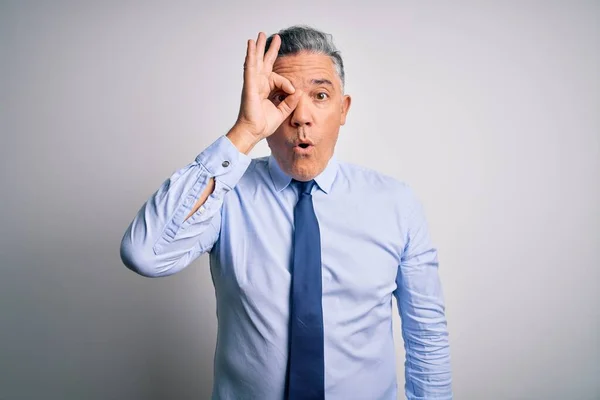 Middle age handsome grey-haired business man wearing elegant shirt and tie doing ok gesture shocked with surprised face, eye looking through fingers. Unbelieving expression.