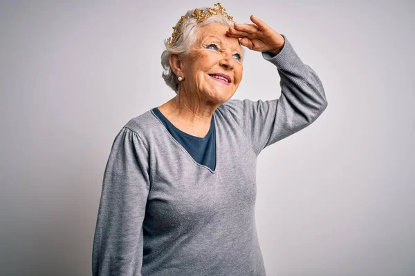 Senior beautiful grey-haired woman wearing golden queen crown over white background very happy and smiling looking far away with hand over head. Searching concept.