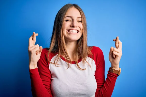 孤立した青の背景にカジュアルなTシャツを着た若い美しい赤毛の女性が希望と目を閉じて笑顔を交差ジェスチャー指 幸運と迷信の概念 — ストック写真