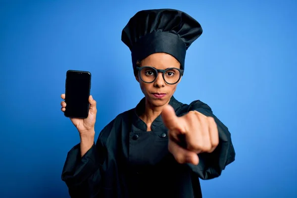 身穿炊具制服的非洲裔美国烘培师女士手持智能手机 屏幕上的手指指向摄像机和你 正面是积极自信的手势 — 图库照片