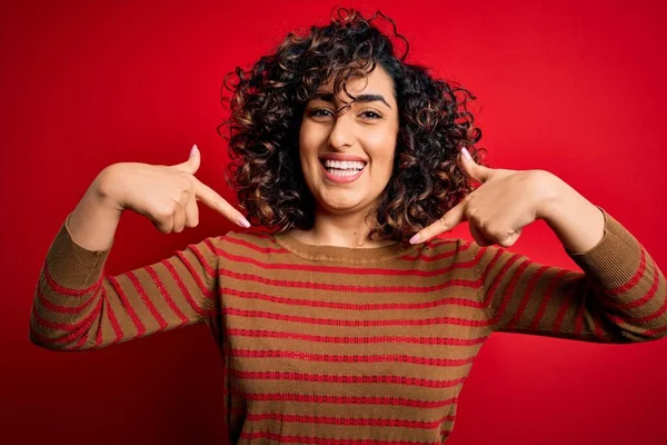 年轻美丽的卷曲阿拉伯女人 身穿宽松的条纹毛衣 站在红色的背景上 满脸笑容 用手指指着自己 自豪而快乐 — 图库照片