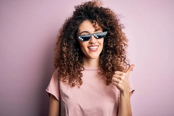 Young beautiful woman with curly hair and piercing wearing funny thug life sunglasses doing happy thumbs up gesture with hand. Approving expression looking at the camera showing success.