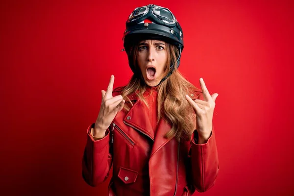 赤い背景に元ヘルメットをかぶっている若い美しいブルネットのオートバイの女性が手を上げて岩のシンボルをやって狂気の表情で叫んでいる 音楽スター 重い概念 — ストック写真