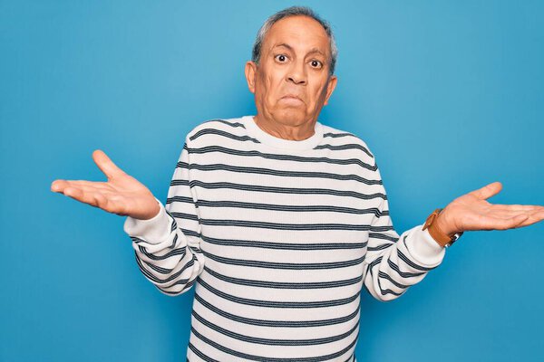 Senior handsome grey-haired man wearing striped sweater over isolated blue background clueless and confused expression with arms and hands raised. Doubt concept.