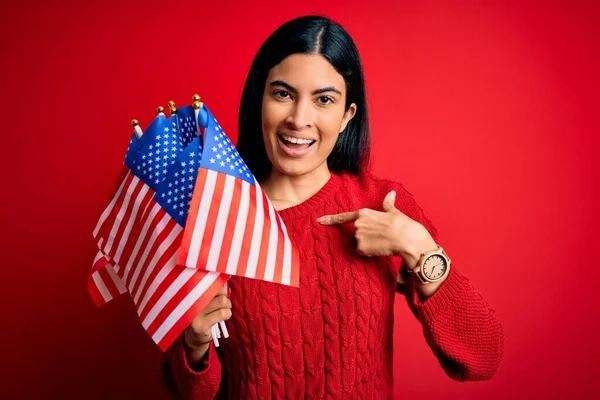 Young beautiful hispanic woman holding United States of American flag at independence day with surprise face pointing finger to himself