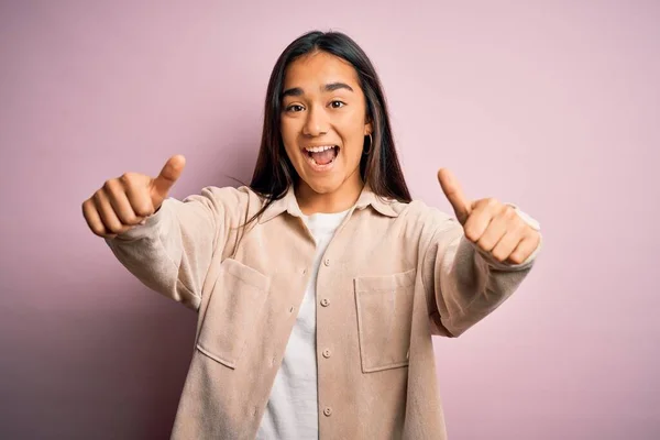 Young beautiful asian woman wearing casual shirt standing over pink background approving doing positive gesture with hand, thumbs up smiling and happy for success. Winner gesture.