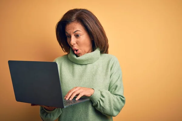中世のブルネット女性使用してコンピュータラップトップ上の黄色の背景怖がってショックで驚きの顔 恐怖と興奮で恐怖表現 — ストック写真