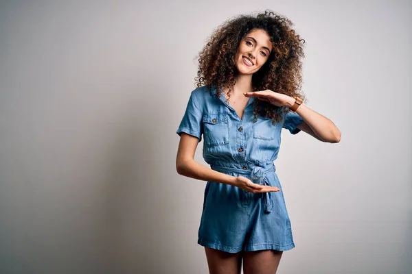 Young beautiful woman with curly hair and piercing wearing casual denim dress gesturing with hands showing big and large size sign, measure symbol. Smiling looking at the camera. Measuring concept.