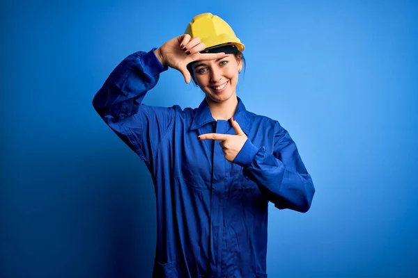 Young beautiful worker woman with blue eyes wearing security helmet and uniform smiling making frame with hands and fingers with happy face. Creativity and photography concept.