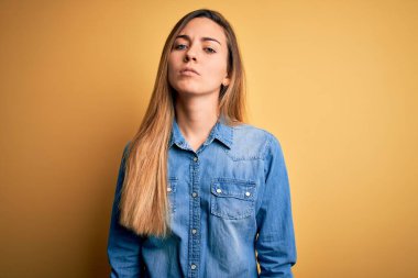 Sarışın, güzel, mavi gözlü, sarı arka planda kot gömlek giyen, yüzünde ciddi bir ifade olan bir kadın. Basit ve doğal bir şekilde kameraya bakıyorsun..