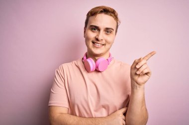 Genç, kızıl saçlı, müzik dinleyen, pembe arka planda kulaklık takan, yüzünde kocaman bir gülümseme olan, eli ve parmağı kameraya bakan genç bir adam..