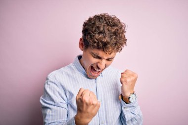 Sarışın, kıvırcık saçlı, beyaz arka planda çizgili tişört giyen genç ve yakışıklı bir adam kolları havada ve gözleri kapalı olarak başarıyı kutluyor ve şaşırıyor. Kazanan konsept.