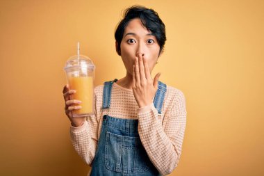 Sağlıklı portakal suyu içen genç, güzel Çinli bir kadın izole edilmiş sarı arka planda ağzı olan bir kadın hata yaptığı için utancından, korku ifadesinden, sessizlikten korkan, gizli bir konseptten korkan bir kadın.