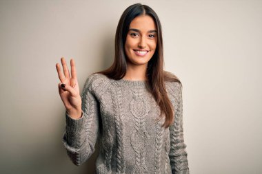 Genç, güzel esmer kadın izole edilmiş beyaz arka plan üzerine günlük süveter giyiyor ve üç numaralı parmaklarıyla işaret ederken kendinden emin ve mutlu bir şekilde gülümsüyor..