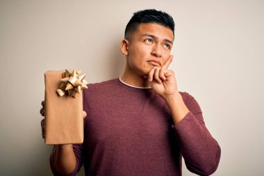 Yakışıklı Latin bir adam, izole edilmiş beyaz arka planda doğum günü hediyesi tutuyor. Ciddi bir yüz. Soru sormayı düşünüyor.