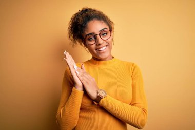 Genç ve güzel Afrikalı Amerikalı kız sarı arka planda süveter ve gözlük takıp alkışlıyor ve mutlu ve neşeli, gururlu elleri alkışlıyor.