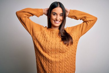 Genç, güzel esmer kadın izole edilmiş beyaz arka planda rahat ve esnek bir süveter giyiyor kolları ve elleri başının arkasında ve boynunun arkasında mutlu bir şekilde gülümsüyor.