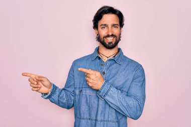 Yakışıklı İspanyol bohem bir adam pembe arka planda hippi tarzı giyiniyor gülümsüyor ve kameraya bakıyor iki eliyle ve parmaklarıyla yandan işaret ediyor..