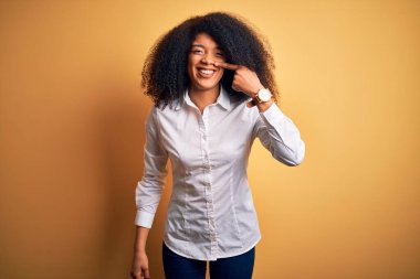 Afro saçlı, sarı arka planda duran, el ve burnu ile işaret eden, neşeli gülümseyen güzel, Afro-Amerikan zarif genç bir kadın. Güzellik konsepti