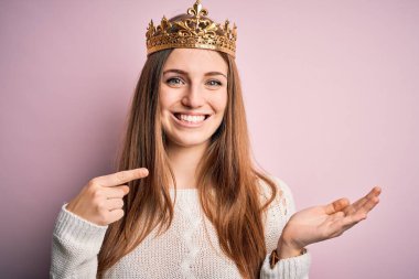 Soyutlanmış pembe arka planda kraliçe tacı takan genç ve güzel bir kızıl kadın kameraya hayret ediyor ve gülümsüyor..