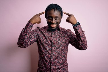 Genç, yakışıklı, Afro-Amerikalı bir adam pembe arka planda dikilmiş gülümsüyor, iki eliyle başını işaret ediyor, harika bir fikir ya da düşünce, iyi bir hafıza.
