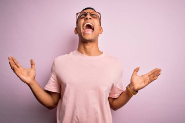 Yakışıklı Afro-Amerikalı adam pembe arka plan üzerine günlük tişört ve gözlük takıyor. Deli gibi bağırıyor, agresif bir ifade ve kollarla bağırıyor. Hayal kırıklığı konsepti.