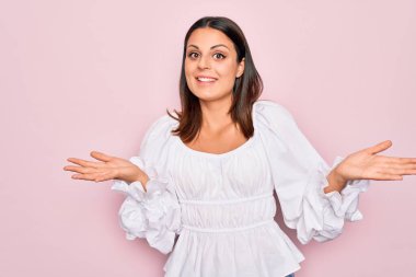 Genç, güzel, esmer, günlük beyaz elbiseler giyen, pembe arka planda duran, kafası karışmış, elleri ve kolları havada bir kadın. Şüphe kavramı.