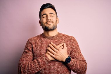 Soyutlanmış pembe arka planda dikilen sıradan bir süveter giyen genç yakışıklı elleri göğsünde, gözleri kapalı ve yüzünde minnet dolu bir jest ile. Sağlık kavramı.