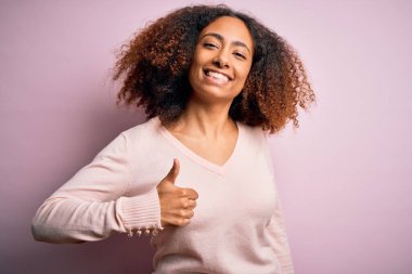 Afro saçlı, pembe arka planda günlük süveter giyen Afro-Amerikalı genç bir kadın elleriyle mutlu bir jest yapıyor. Kameraya bakıp başarıyı gösteren ifade onaylanıyor.