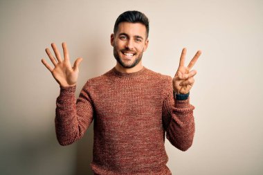 İzole edilmiş beyaz arka plan üzerinde duran sıradan süveter giyen genç yakışıklı adam yedinci parmaklarını gösterirken kendinden emin ve mutlu bir şekilde gülümsüyor..