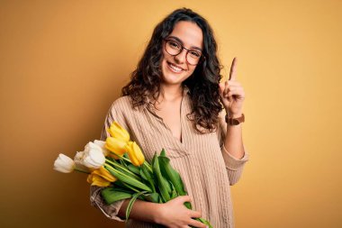 Sarı lalelerden bir buket buket tutan genç ve güzel romantik bir kadın. Şaşırmış ve şaşırmış bir şekilde gülümsüyor. Parmaklarıyla işaret ediyor ve kollarını kaldırıyor..