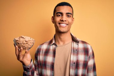 Genç ve yakışıklı Afro-Amerikalı adam elinde kaseyle sağlıklı mısır gevrekleri ve mutlu bir yüzle ayakta duruyor ve gülümsüyor dişlerini gösteren bir gülümsemeyle