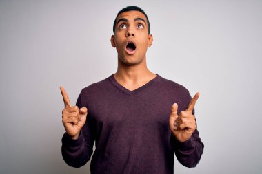 Beyaz arka plan üzerine gündelik süveter giyen genç ve yakışıklı Afro-Amerikalı adam şaşırmış ve şaşırmış bir şekilde yukarı bakıyor ve parmaklarıyla işaret ediyor ve kollarını kaldırıyor..