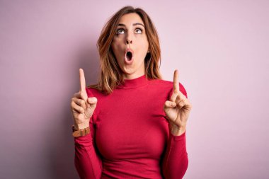 Genç, güzel, esmer bir kadın pembe arka planda beliren gündelik bir süveter giyiyor şaşkın şaşkın yukarı bakıyor ve parmaklarıyla işaret ediyor ve kollarını kaldırıyor..