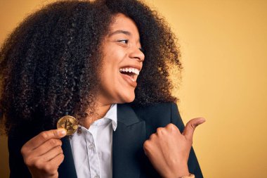 Afro saçlı, sarı arka planda bitcoin tutan ve gülümseyen bir gülümsemeyle baş parmağıyla gösteren Afro-Amerikalı genç bir iş kadını.