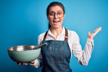 Mavi gözlü genç, güzel fırıncı kadın tencere ve çırpıcıyla izole arka planda mutlu ve heyecanlı, kazanan yüz ifadesi zaferi büyük bir gülümsemeyle kutluyor ve ellerini kaldırıyor.