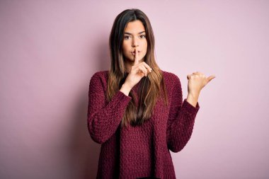 İzole edilmiş pembe arka plan üzerine sıradan süveter giyen genç ve güzel bir kız sessiz olmak istiyor. Parmakları dudaklarında, elleri yana doğru işaret ederek. Sessizlik ve gizli konsept.