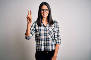 Mavi gözlü genç esmer kadın beyaz arka planda günlük gömlek ve gözlük takıyor ve parmaklarını iki numarayı gösterirken kendinden emin ve mutlu bir şekilde gülümsüyor..