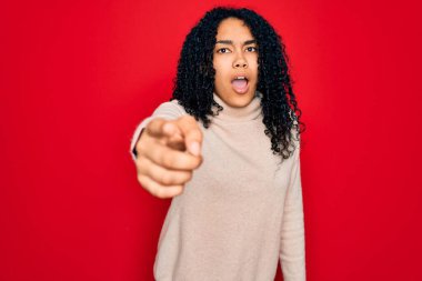 Genç Afro-Amerikan kıvırcık saçlı kadın kırmızı arka plan üzerine balıkçı yaka kazak giyiyor. Kameraya kızgın ve sinirli olduğunu gösteriyor.