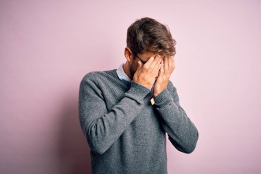 Sakallı, gözlüklü ve süveterli genç bir adam pembe arka planda dikiliyor. Ağlarken suratını ellerle kapatıyor. Depresyon kavramı.