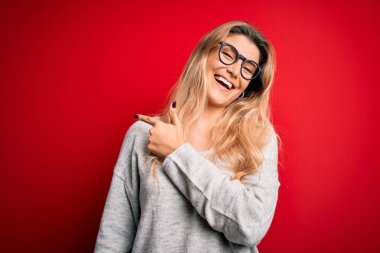 Genç, sarışın, süveter ve gözlük takan güzel bir kadın izole edilmiş kırmızı arka planda neşeli, yüzünde mutlu ve doğal bir ifadeyle el ve parmak ile gülümseyen bir gülümseme var.