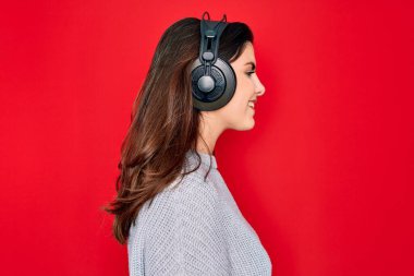 Modern kulaklık takan genç ve güzel bir kız kırmızı arka planda müzik dinliyor. Yan tarafa bakıyor, rahat profil pozu veriyor. Kendine güvenen bir gülümsemesi var..