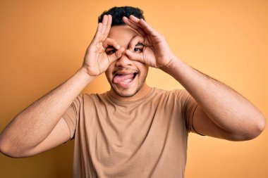 İzole edilmiş sarı arka plan üzerinde duran sıradan tişört giyen genç yakışıklı bir adam dürbün gibi dil çıkartıyor, gözler parmaklarından bakıyor. Çılgın ifade.