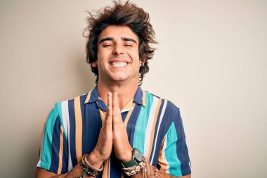 Yaz tatilinde olan genç bir adam, renkli tişörtler giyerek izole edilmiş beyaz arka planda duruyor. Elleriyle yalvarıyor ve dua ediyor. Yüzünde umut ifadesi var. Çok duygusal ve endişeli. Yalvarıyorum..