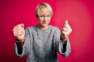 Modern kısa saçlı genç bir kadın pembe arka plan üzerine gündelik süveter giyiyor kızgın ve kızgın yumruklarını kaldırıyor öfkeyle bağırırken sinirli ve öfkeli. Öfke ve agresif kavram.