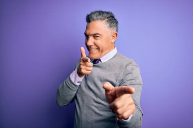 Orta yaşlı, yakışıklı, gri saçlı, mor arka planda zarif bir süveter giyen, mutlu ve komik yüzlü parmakları kameraya gösteren bir adam. İyi enerji ve titreşimler.
