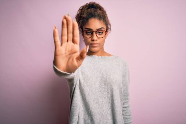 Genç ve güzel Afro-Amerikalı kız pembe arka planda süveter ve gözlük takıyor ve avuç içiyle şarkı söylemeyi kesiyor. Yüzünde olumsuz ve ciddi bir jest olan uyarı ifadesi.